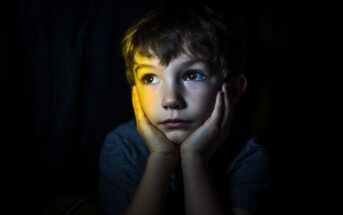 A young boy rests his face in his hands, gazing upward thoughtfully. He is illuminated by a soft, yellow light in an otherwise dark surrounding, highlighting his expressive eyes and pensive expression.