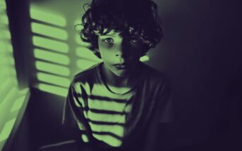 A young boy with curly hair sits in a dimly lit room, illuminated by green light streaming through window blinds, creating striped shadows across his face and shirt.