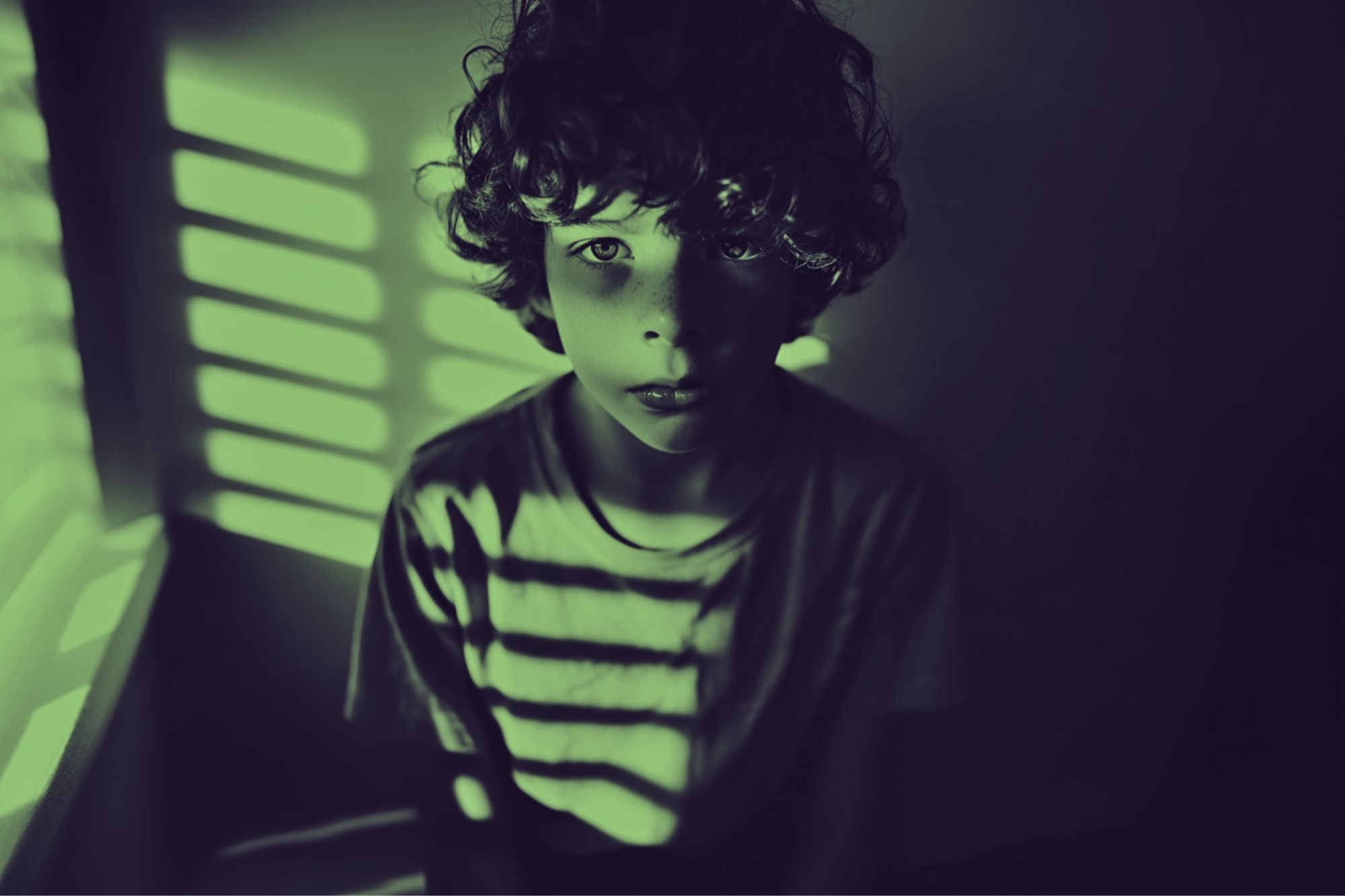 A young boy with curly hair sits in a dimly lit room, illuminated by green light streaming through window blinds, creating striped shadows across his face and shirt.