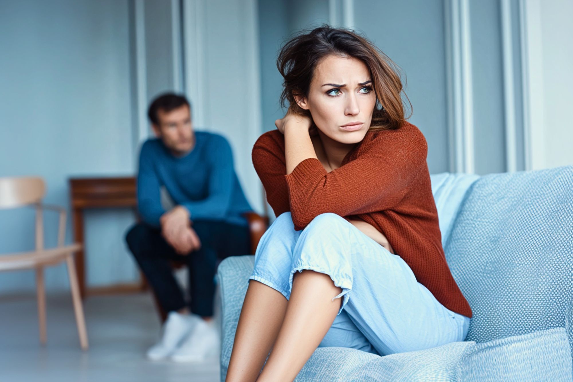 A woman sits on a couch looking upset with her arms around her knees, while a man sits in the background, both appearing distant and troubled in a modern living room.