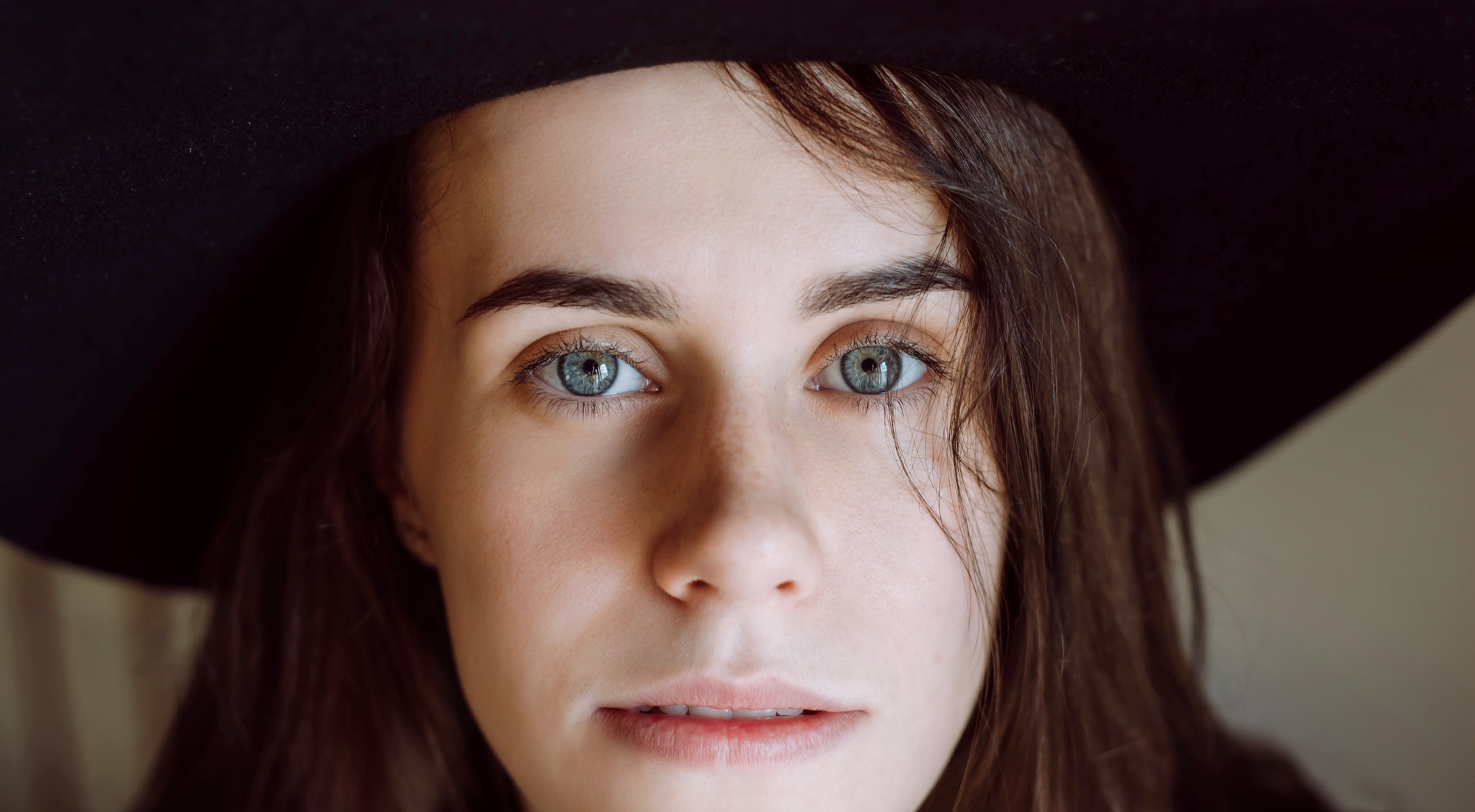 A close-up of a person with long brown hair and blue eyes, wearing a wide-brimmed black hat. They have a neutral expression, and one side of their face is partially covered by hair. The background is softly blurred.