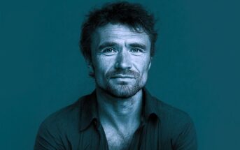 A man with short, wavy hair and a beard looks directly at the camera with a neutral expression. The image has a blue monochrome filter and a textured, sketch-like effect. He wears an open-collared shirt.