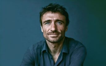 A man with tousled brown hair, blue eyes, and a slight beard wears a dark blue shirt, sitting against a plain blue background, looking directly at the camera with a neutral expression.
