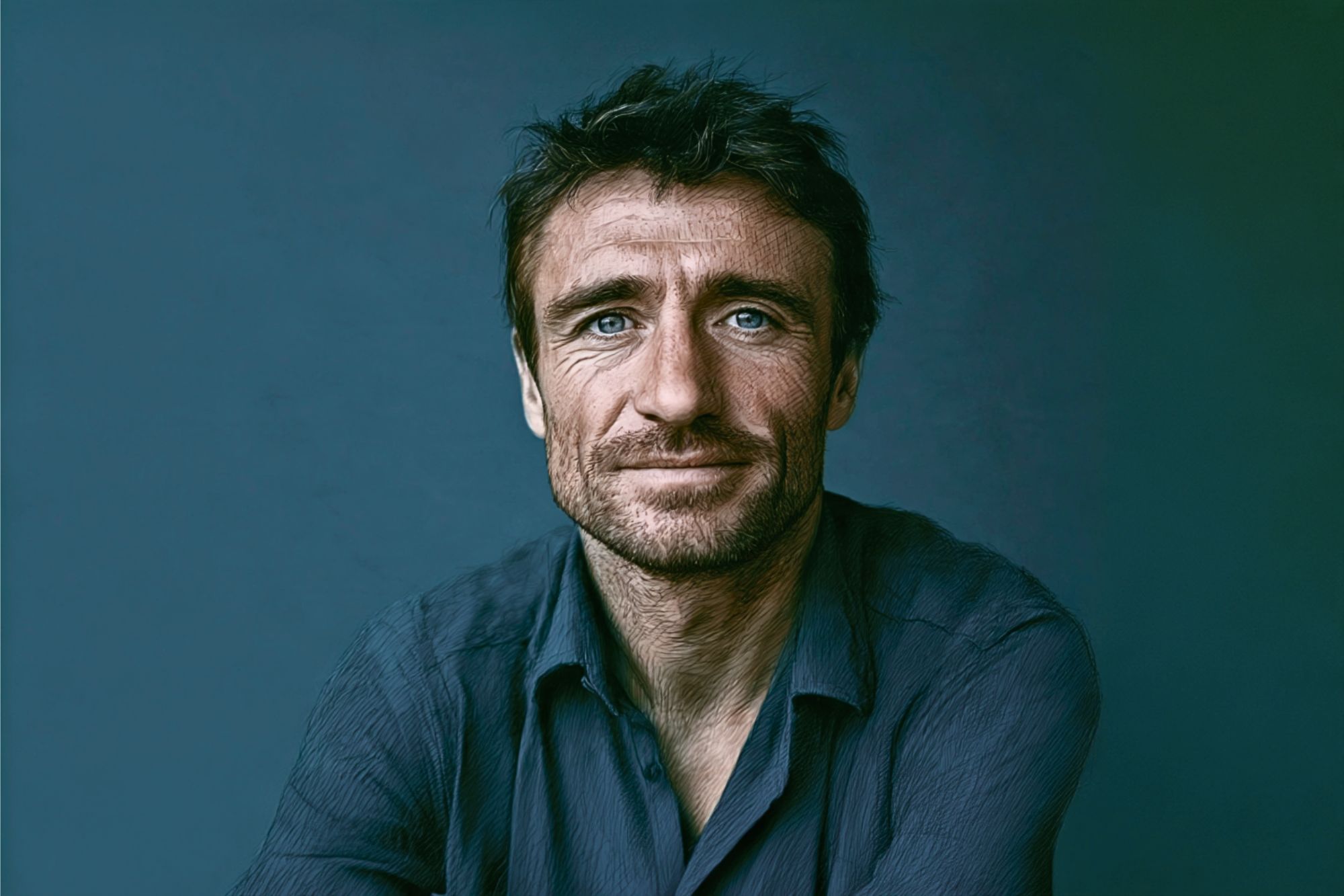 A man with tousled brown hair, blue eyes, and a slight beard wears a dark blue shirt, sitting against a plain blue background, looking directly at the camera with a neutral expression.