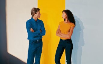 A man and woman stand facing each other with arms crossed, against a wall with blue, yellow, and white vertical stripes. They appear to be having a serious conversation.