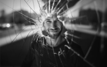 A man with a beard smiles through a jagged hole in a cracked glass window, with blurred buildings in the background. The image is in black and white.