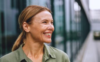 A woman with light brown hair tied back smiles while looking to the side. She is wearing a green blouse and standing outdoors with a blurred modern building in the background.