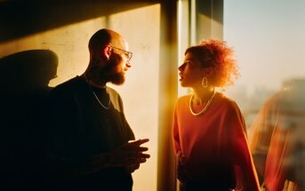 A man with tattoos and glasses talks to a woman with curly hair and hoop earrings as warm sunlight streams through a window, casting dramatic shadows on the wall.