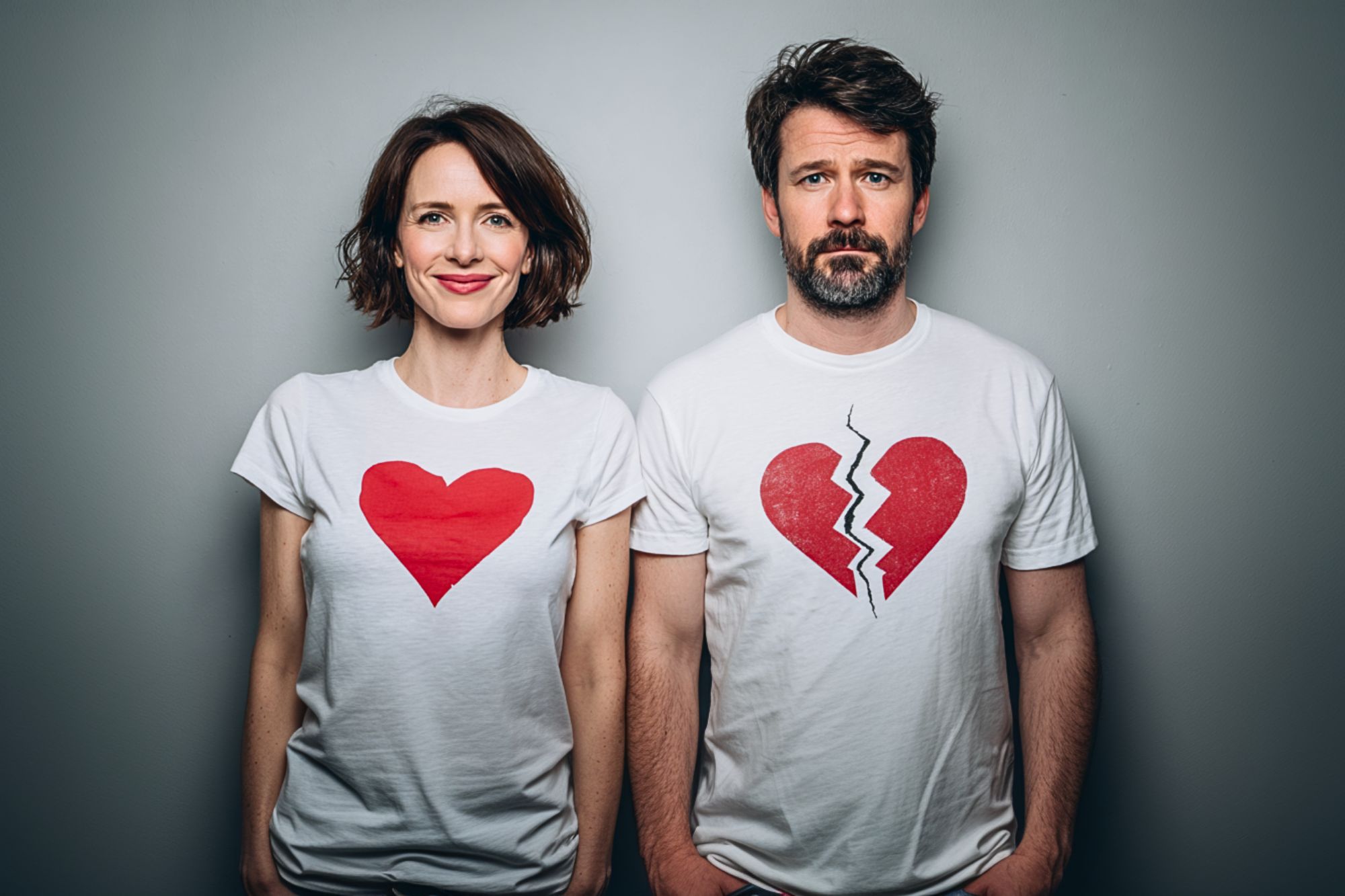 A woman and a man stand side by side against a gray wall. The woman is smiling and wears a shirt with a red heart, while the man looks serious and wears a shirt with a red broken heart.