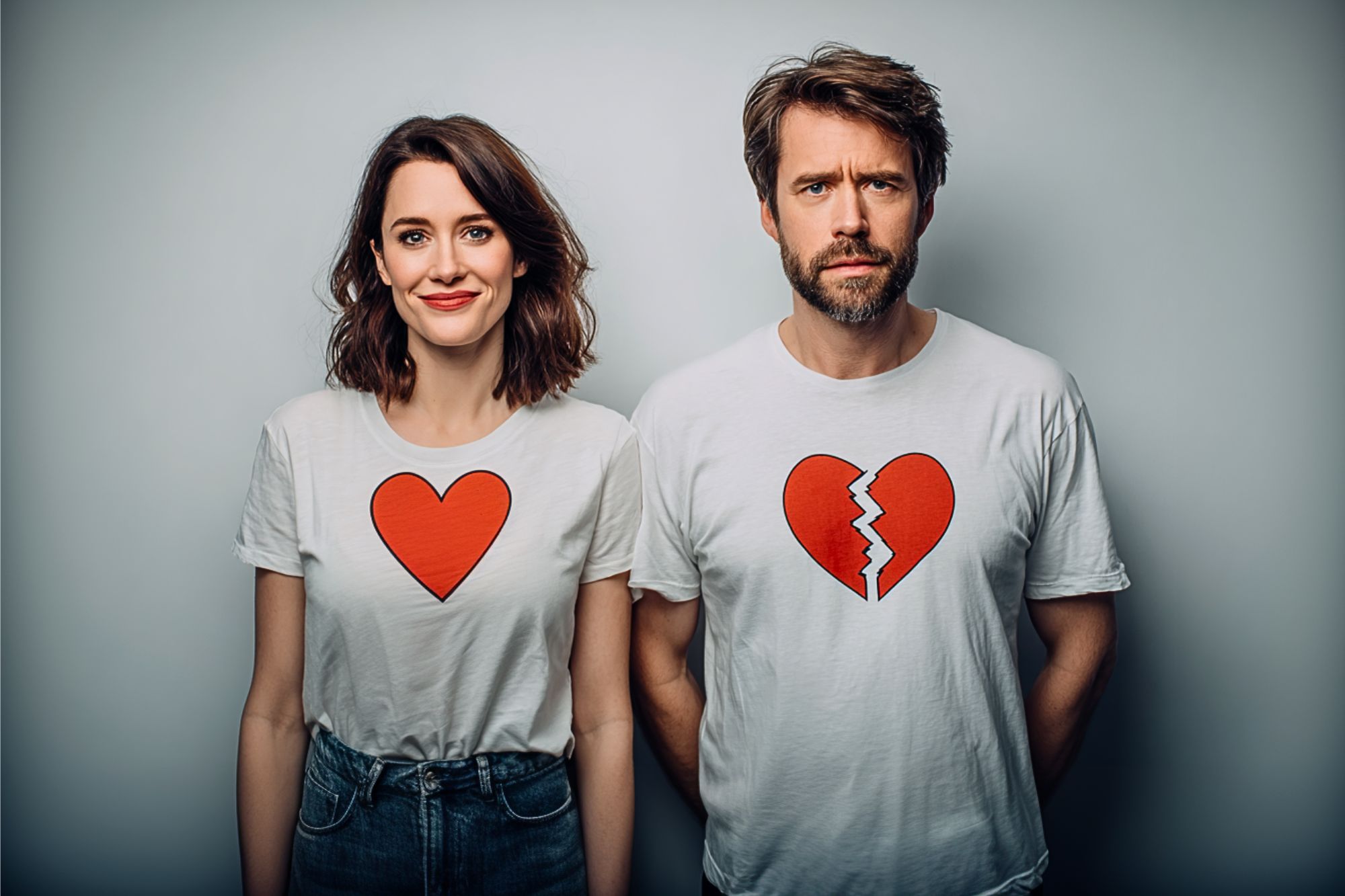 A woman smiles wearing a white t-shirt with a red heart, while a man beside her looks serious in a matching shirt with a red broken heart. Both stand against a plain gray background.