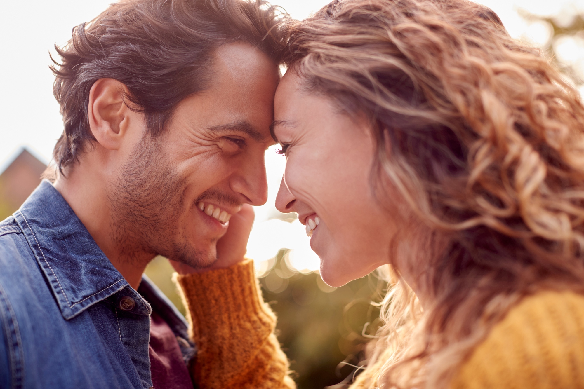 A smiling couple stands face-to-face, leaning their foreheads together. The man is wearing a denim jacket, and the woman is in a yellow sweater. They are outdoors with a blurred background, suggesting a sunny day.