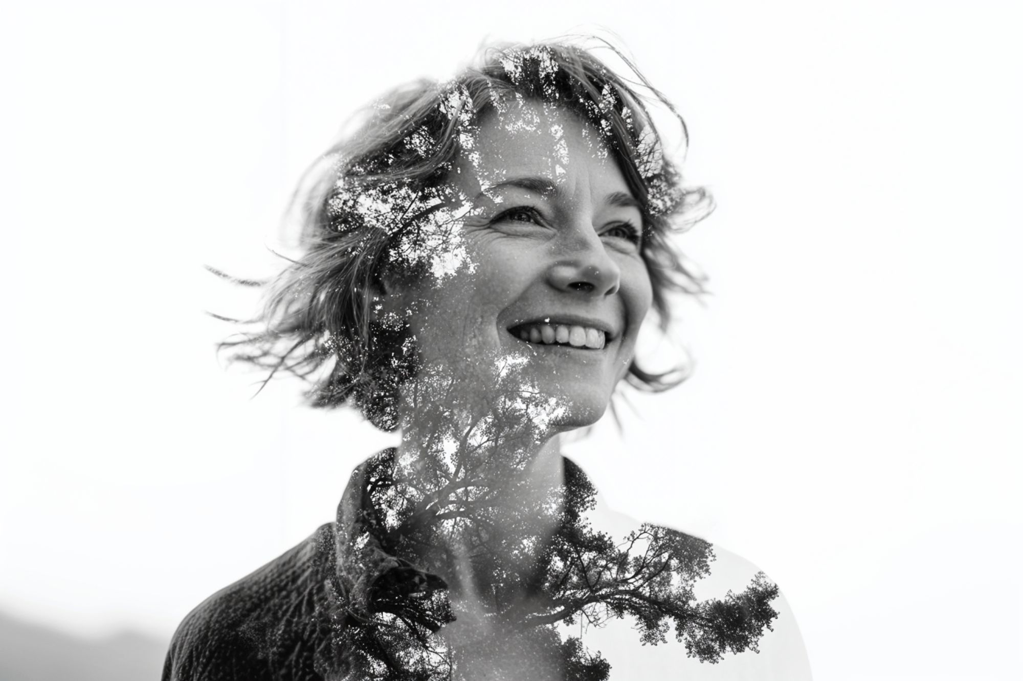 A black and white double exposure image of a smiling woman with short hair, blended with the branches and leaves of a tree, creating a harmonious natural effect.