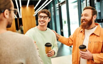 Three men are standing indoors, smiling and talking. Two of them are holding coffee cups. The man on the right, wearing an orange jacket and with tattoos, is patting the shoulder of another man in glasses and a light green sweater.