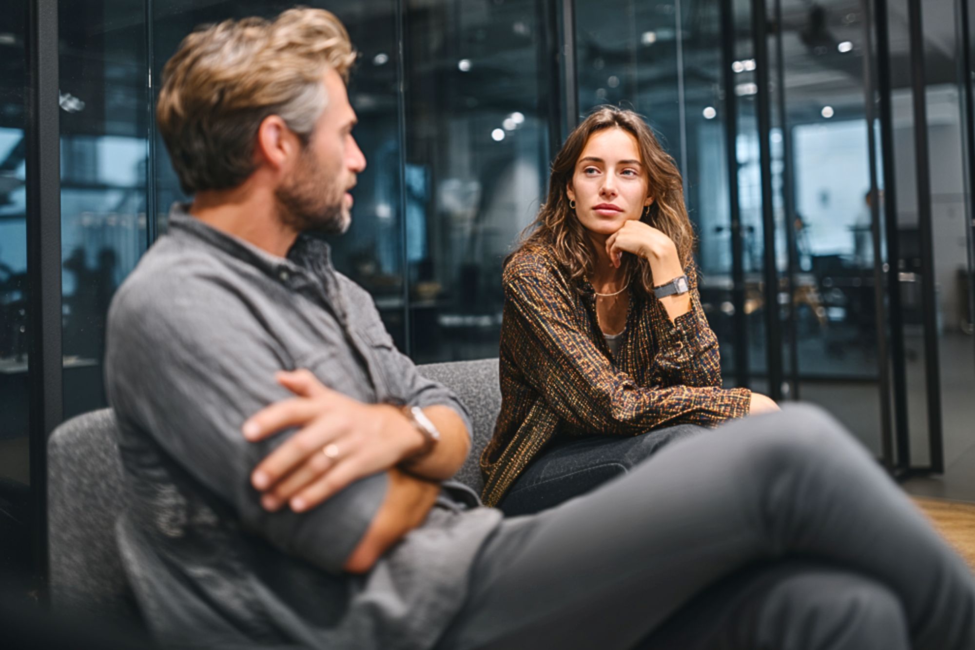 A man and a woman sit on a sofa in a modern office, engaged in a serious conversation. The woman looks attentively at the man, who has his arms crossed. Glass walls and office furniture are visible in the background.