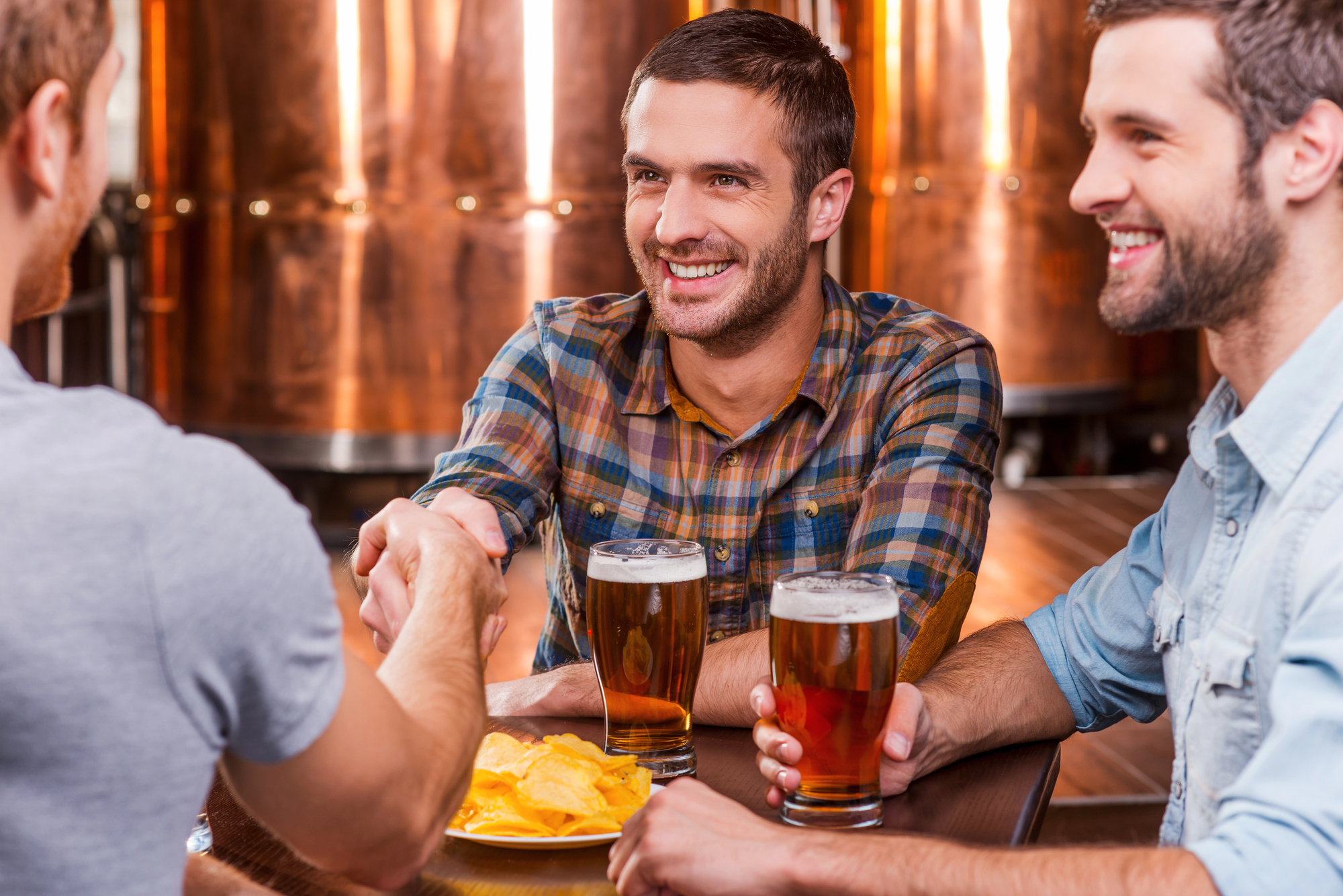 Three men sitting at a brewery table, holding glasses of beer. One man in a plaid shirt is shaking hands with another man whose back is to the camera. A bowl of chips is on the table. They are smiling with copper brewing tanks in the background.