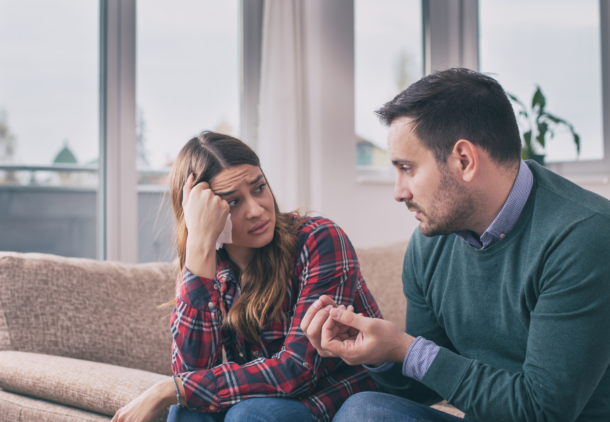 A man and a woman are sitting on a couch in a living room. The man is gesturing with his hands, explaining something, while the woman looks concerned and rests her head on her hand. Large windows are in the background.