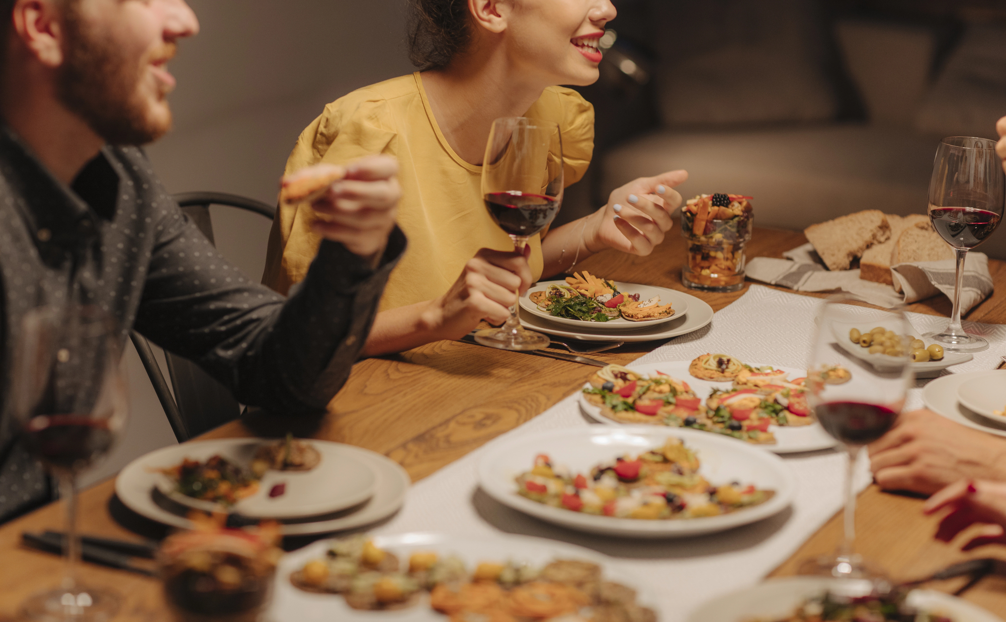 A group of people sitting around a wooden table enjoying a meal and drinks. Various dishes, including salads and appetizers, are spread across the table, along with glasses of red wine. One person in a yellow shirt is smiling and holding a fork.