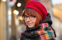 A person with short brown hair wears glasses, a red beret, and a colorful plaid scarf, smiling softly outdoors with blurred lights and buildings in the background.