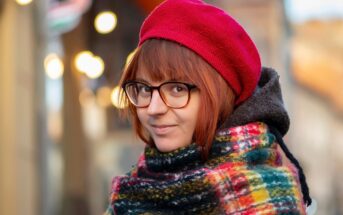 A person with short brown hair wears glasses, a red beret, and a colorful plaid scarf, smiling softly outdoors with blurred lights and buildings in the background.