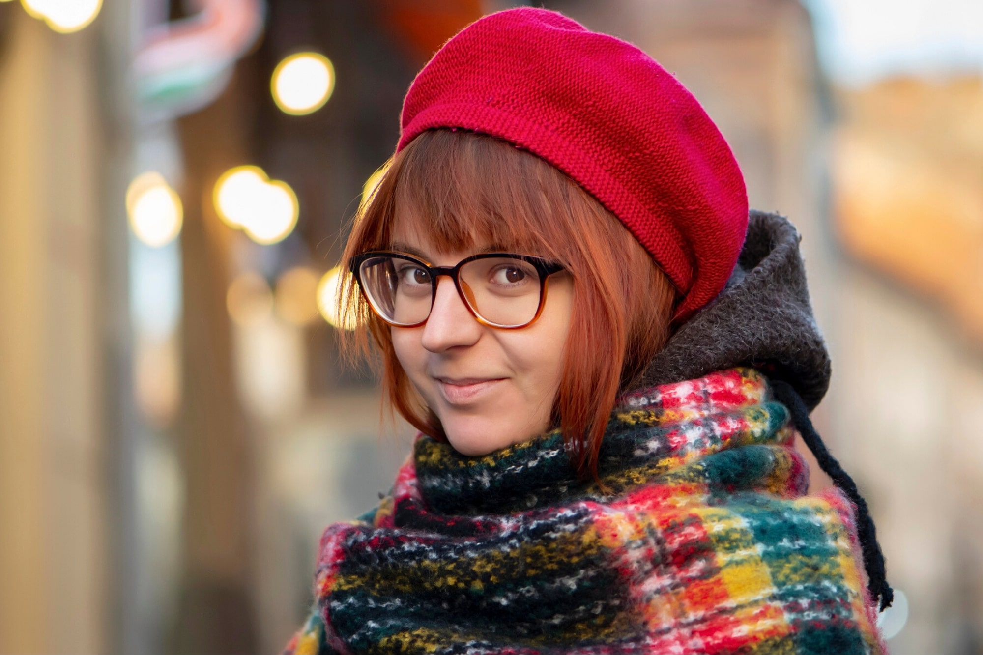 A person with short brown hair wears glasses, a red beret, and a colorful plaid scarf, smiling softly outdoors with blurred lights and buildings in the background.