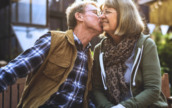 An elderly couple sits closely on a bench outdoors. The man, wearing a plaid shirt and brown vest, kisses the woman on the cheek. She wears glasses, a green jacket, and a scarf, smiling warmly. Sunlight and triangular banners are in the background.