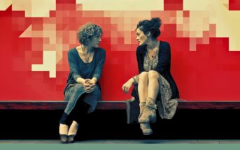 Two women sit on a bench against a red and pixelated background, facing each other and engaged in conversation. Both wear casual dresses and boots, with one woman crossing her legs and the other clasping her hands.