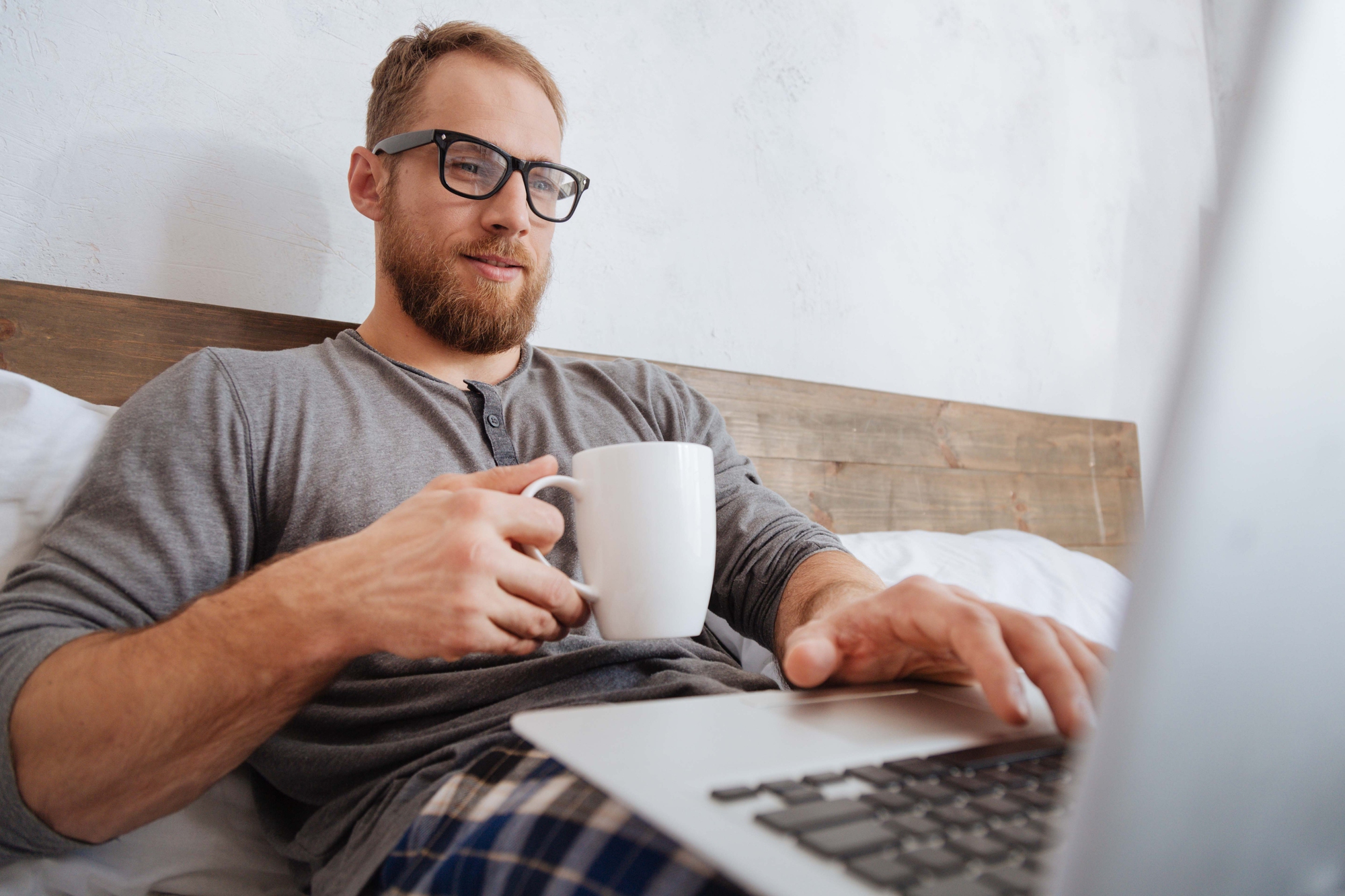 A man with glasses and a beard is sitting on a bed, holding a white mug in one hand and typing on a laptop with the other. He is wearing a gray shirt and checkered pajama pants, and he appears relaxed.