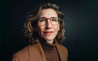 A woman with wavy hair and glasses, wearing a brown turtleneck and blazer, stands against a dark background, smiling softly at the camera.