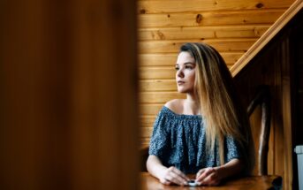 A woman with long hair sits at a wooden table, looking thoughtfully to the side. She is wearing an off-the-shoulder blue patterned top. The background features wooden paneled walls.