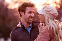 A couple smiling lovingly at each other in a warm, outdoor setting. The man is wearing a dark sweater, and the woman is in a light hat and sweater. The background is softly blurred, suggesting a sunset or sunrise.