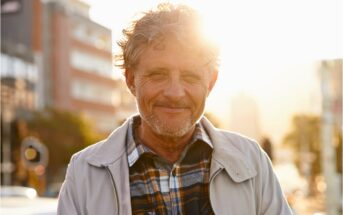 A man with curly hair and a beard smiles while standing outdoors in a city. He is wearing a plaid shirt and light jacket. The sun is setting behind him, creating a warm glow. Urban buildings are visible in the background.