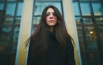 A young woman with long brown hair and a neutral expression stands in front of a building with large glass windows, wearing a dark coat. The background shows reflections of other structures.