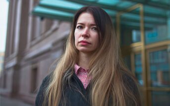 A woman with long, light brown hair stands outside a building with glass doors, wearing a black coat over a red and white checkered shirt. She has a serious expression on her face.