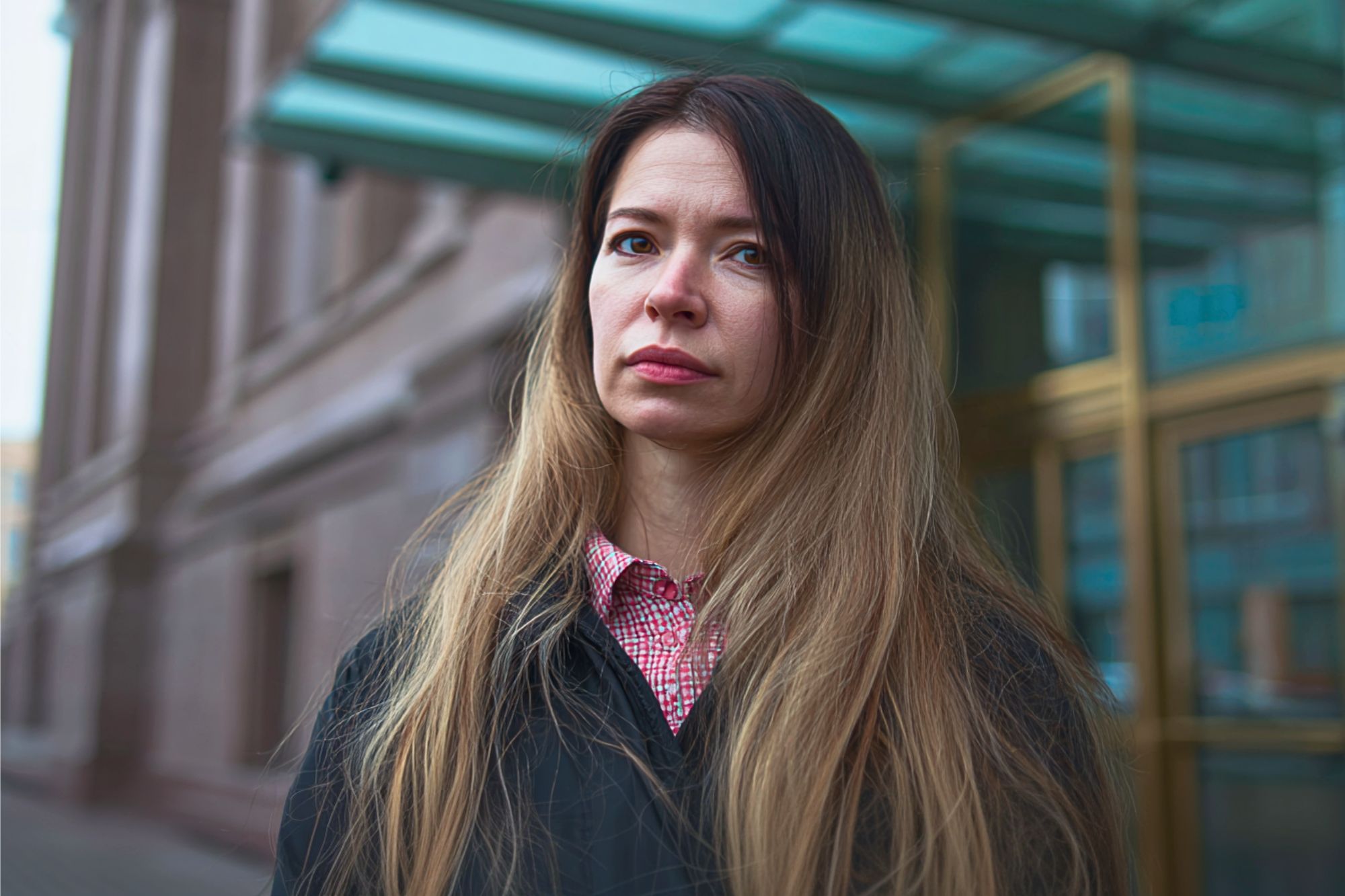 A woman with long, light brown hair stands outside a building with glass doors, wearing a black coat over a red and white checkered shirt. She has a serious expression on her face.