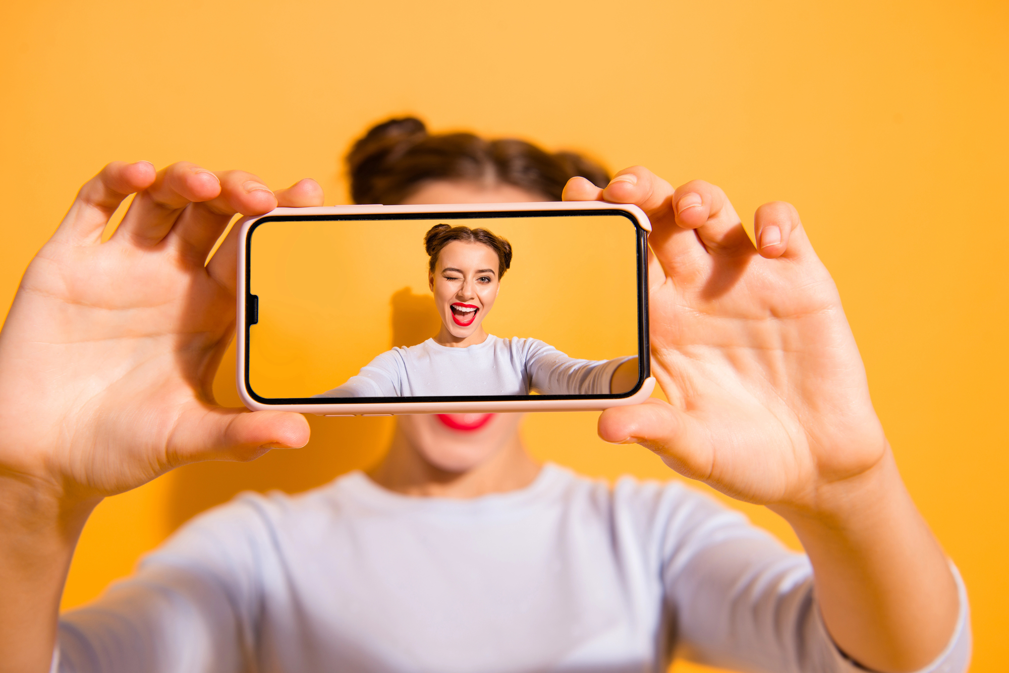 A person holds a smartphone in front of their face, capturing a selfie. The phone displays their excited expression with a wide smile. The background is a vibrant yellow, and the person wears a light shirt with their hair styled in two buns.