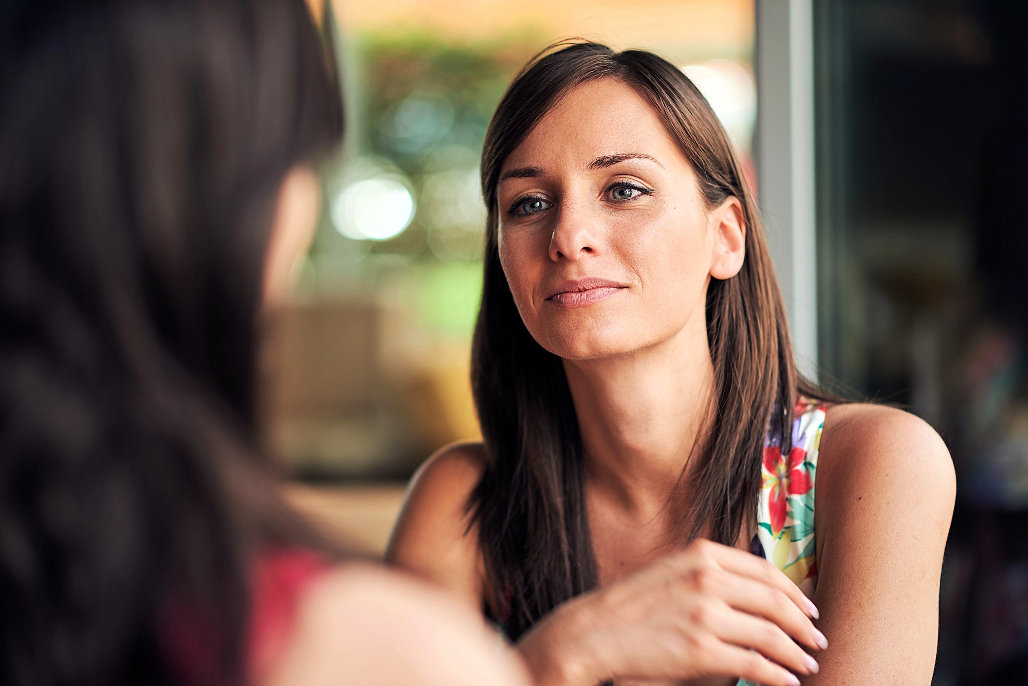 A woman with long brown hair is sitting indoors, wearing a colorful floral top. She is gazing thoughtfully at someone out of frame, with a soft, relaxed expression. The background is softly blurred, suggesting a bright, casual setting.
