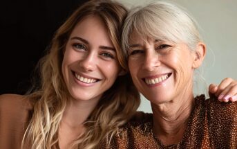 A young woman and an older woman, both smiling, sit close together with their heads touching. The younger woman has long blonde hair, and the older woman has gray hair in a ponytail.