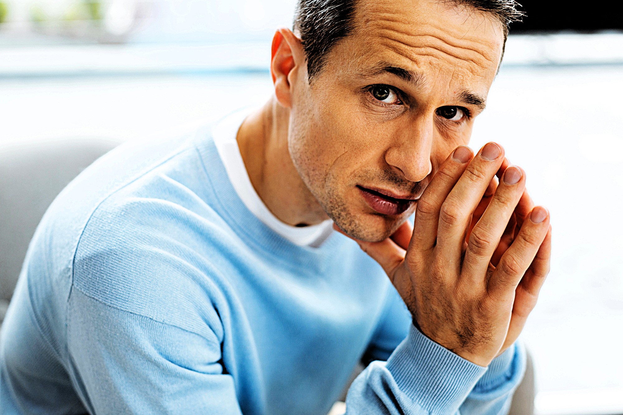 A man in a light blue sweater rests his chin on his hands, looking intently at the camera with a thoughtful expression. The background is blurred, emphasizing the man’s face and contemplative pose.