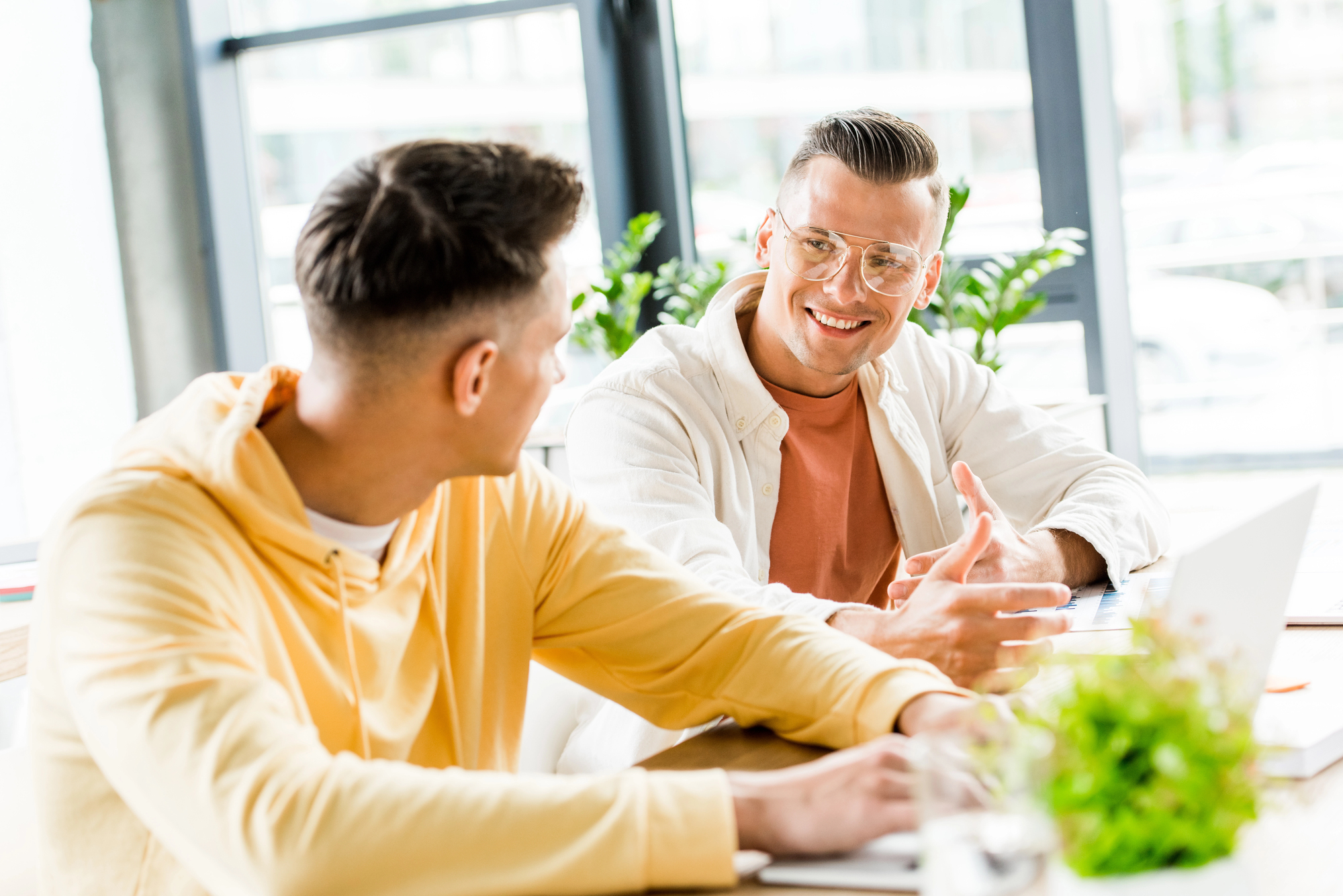 Two men are sitting at a table in a bright room, engaged in conversation. One is wearing glasses and an orange shirt under a light jacket, and the other is in a yellow hoodie. A laptop and a plant are on the table.