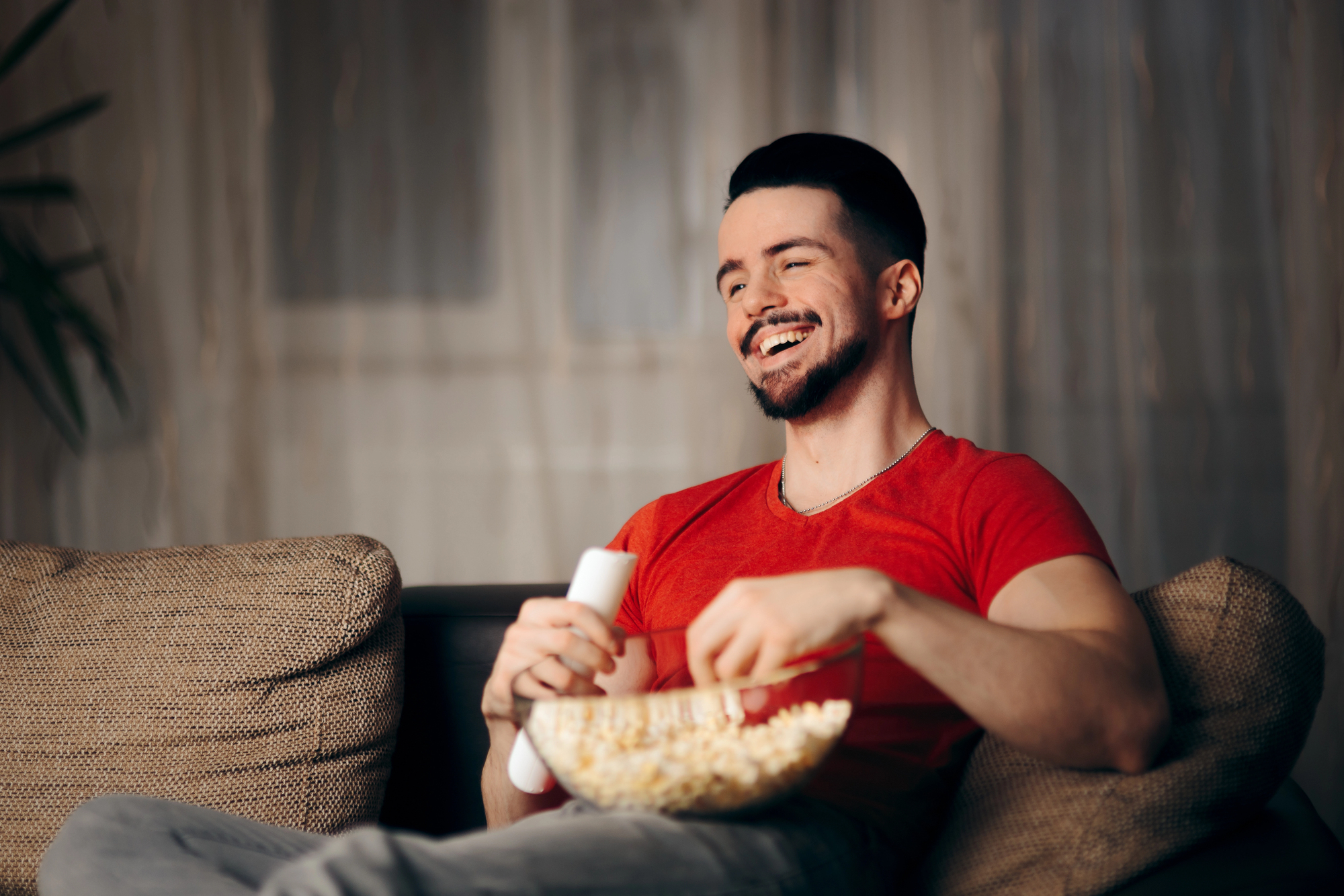 A man in a red shirt sits on a couch, holding a remote control and a bowl of popcorn. He is smiling and appears to be enjoying a show or movie. The background is softly lit, creating a cozy atmosphere.