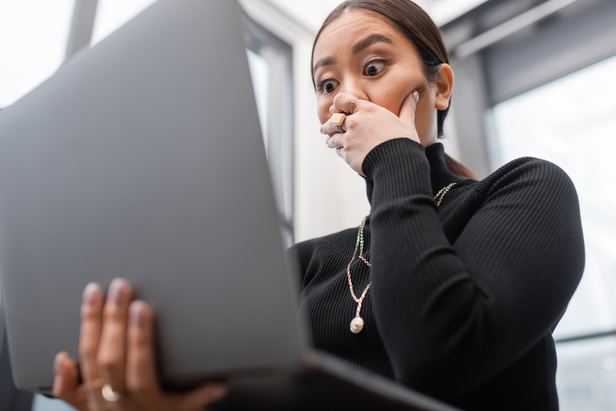 A surprised woman with long hair and a black turtleneck holds a laptop, covering her mouth with her hand, standing in a bright room with large windows.