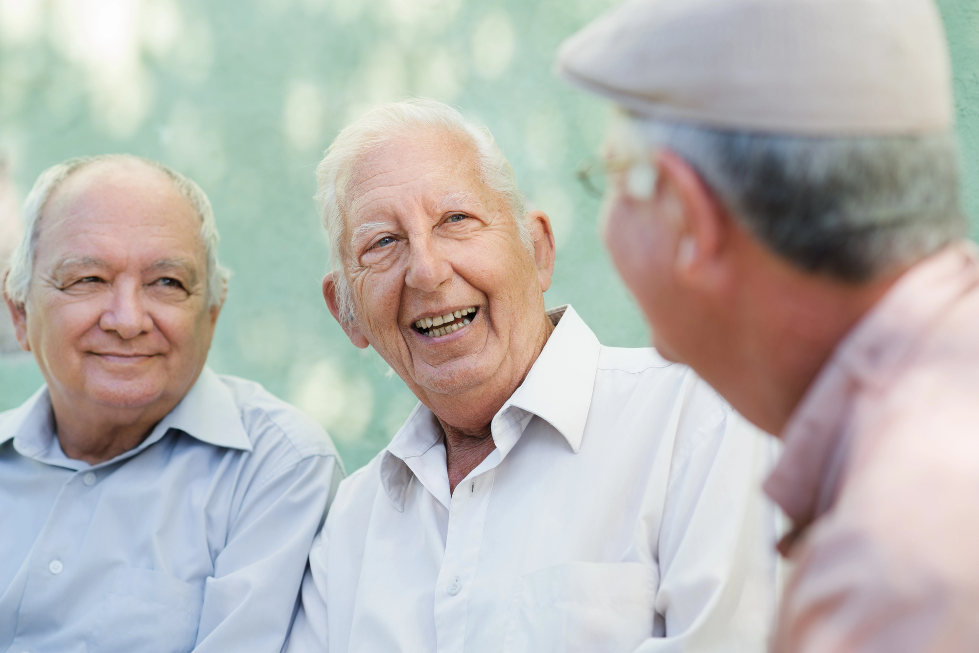 Three elderly men are sitting and laughing together outdoors. The man in the middle is smiling widely, wearing a white shirt. The other two, also in light-colored shirts, are looking at him with happy expressions. The background is a soft green.
