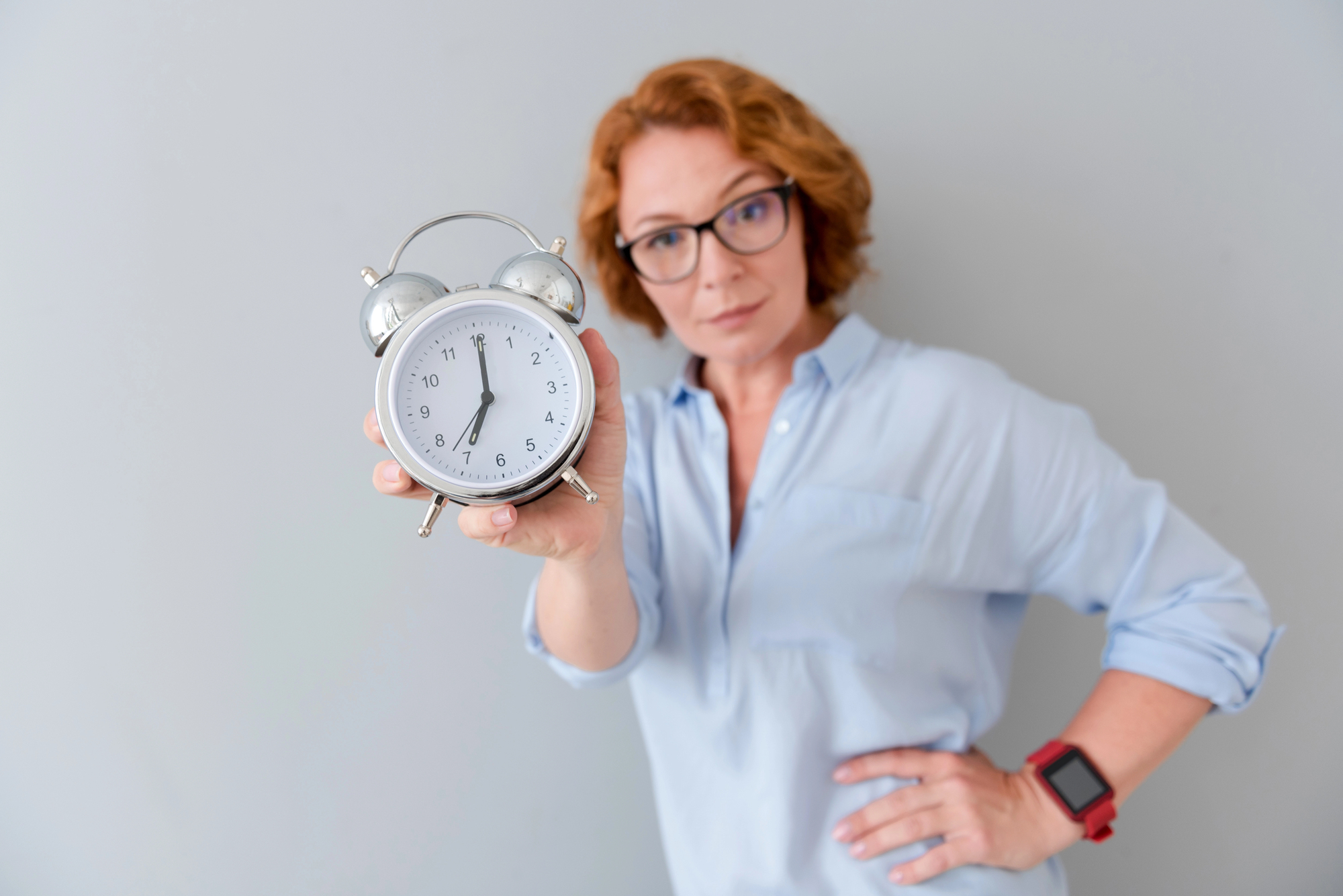 A woman with short reddish hair and glasses holds a silver alarm clock showing 7:00. She wears a light blue shirt and a smartwatch on her wrist, standing against a plain gray background, looking directly at the camera.