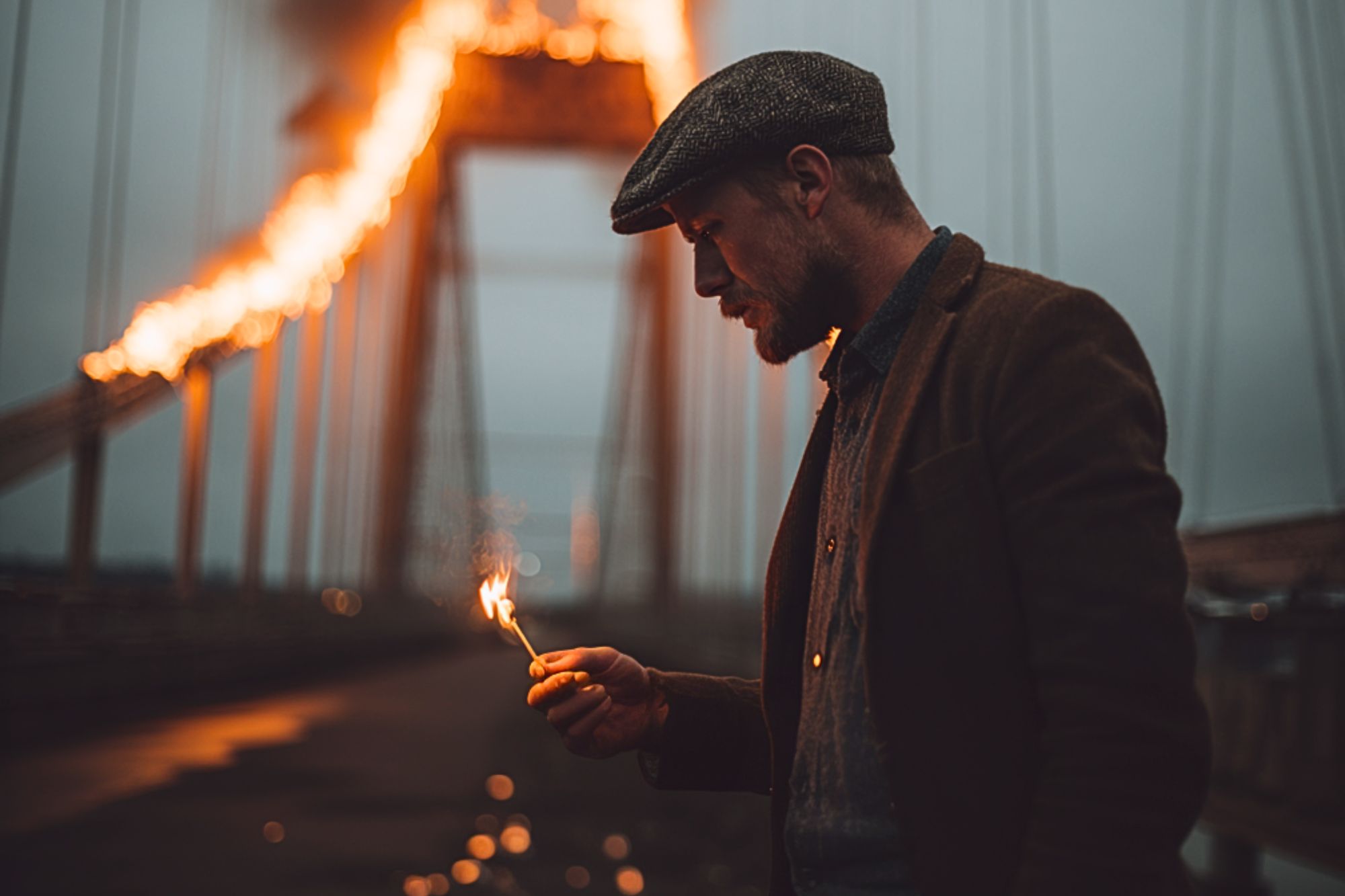 A man in a flat cap and jacket holds a lit match on a bridge at dusk, while large flames burn dramatically along the bridge’s cables in the background.