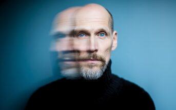 A bald man with a beard and blue eyes in a black turtleneck is shown against a blue background, with a blurred double exposure effect creating a duplicate of his face to the left.