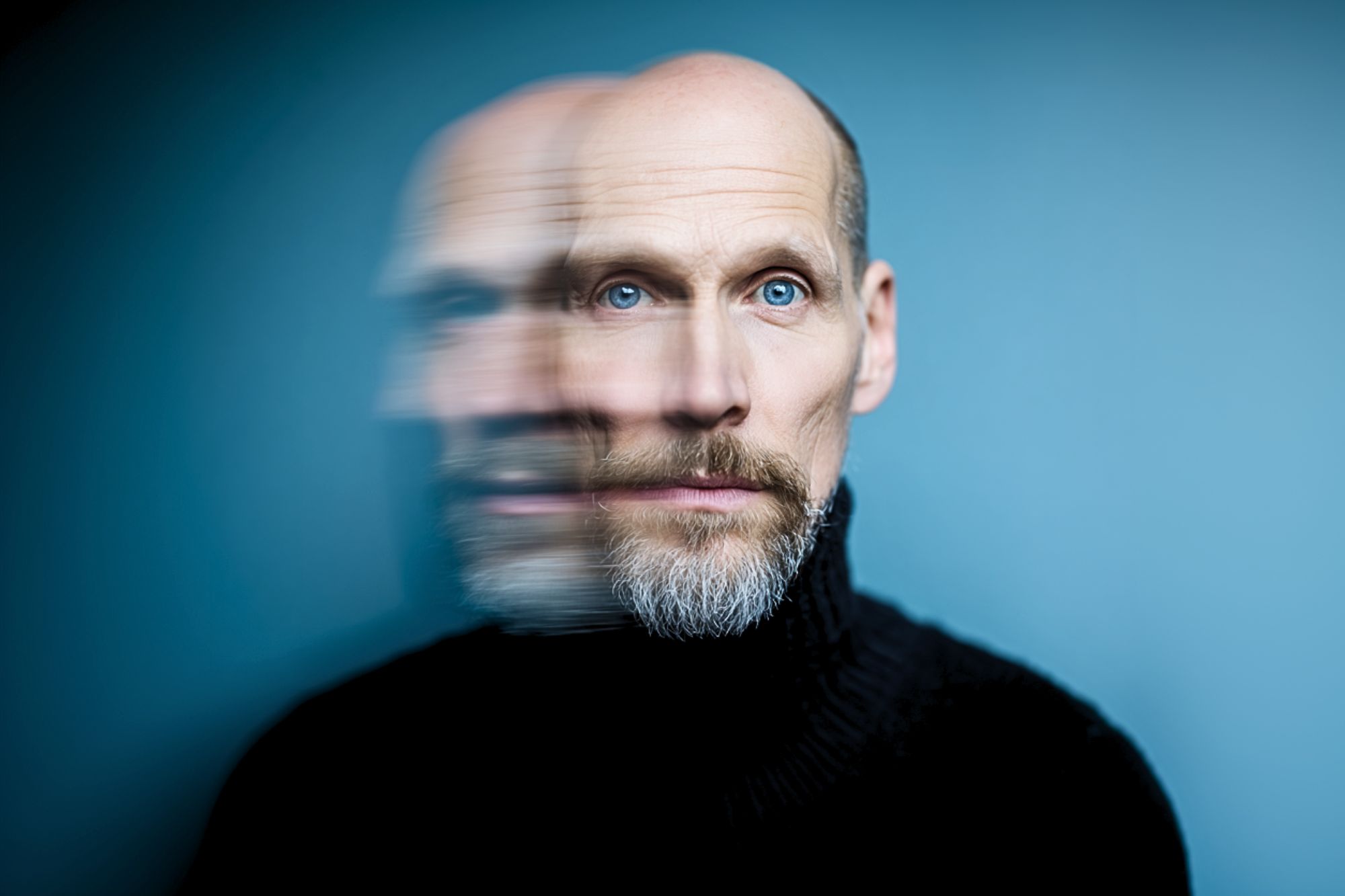 A bald man with a beard and blue eyes in a black turtleneck is shown against a blue background, with a blurred double exposure effect creating a duplicate of his face to the left.