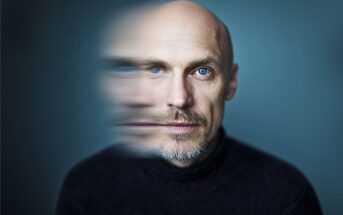 A bald man with blue eyes and a short beard is wearing a dark turtleneck. Half of his face is clear, while the other half is blurred, creating a surreal, double-exposure effect against a blue background.