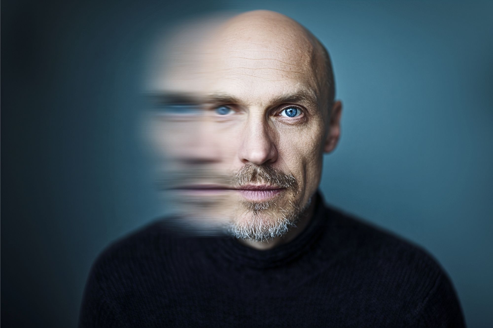 A bald man with blue eyes and a short beard is wearing a dark turtleneck. Half of his face is clear, while the other half is blurred, creating a surreal, double-exposure effect against a blue background.