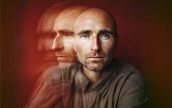 A man with blue eyes and a trimmed beard sits against a reddish background. His face appears twice—the main face in focus and a blurred duplicate to the left, creating a double exposure effect.