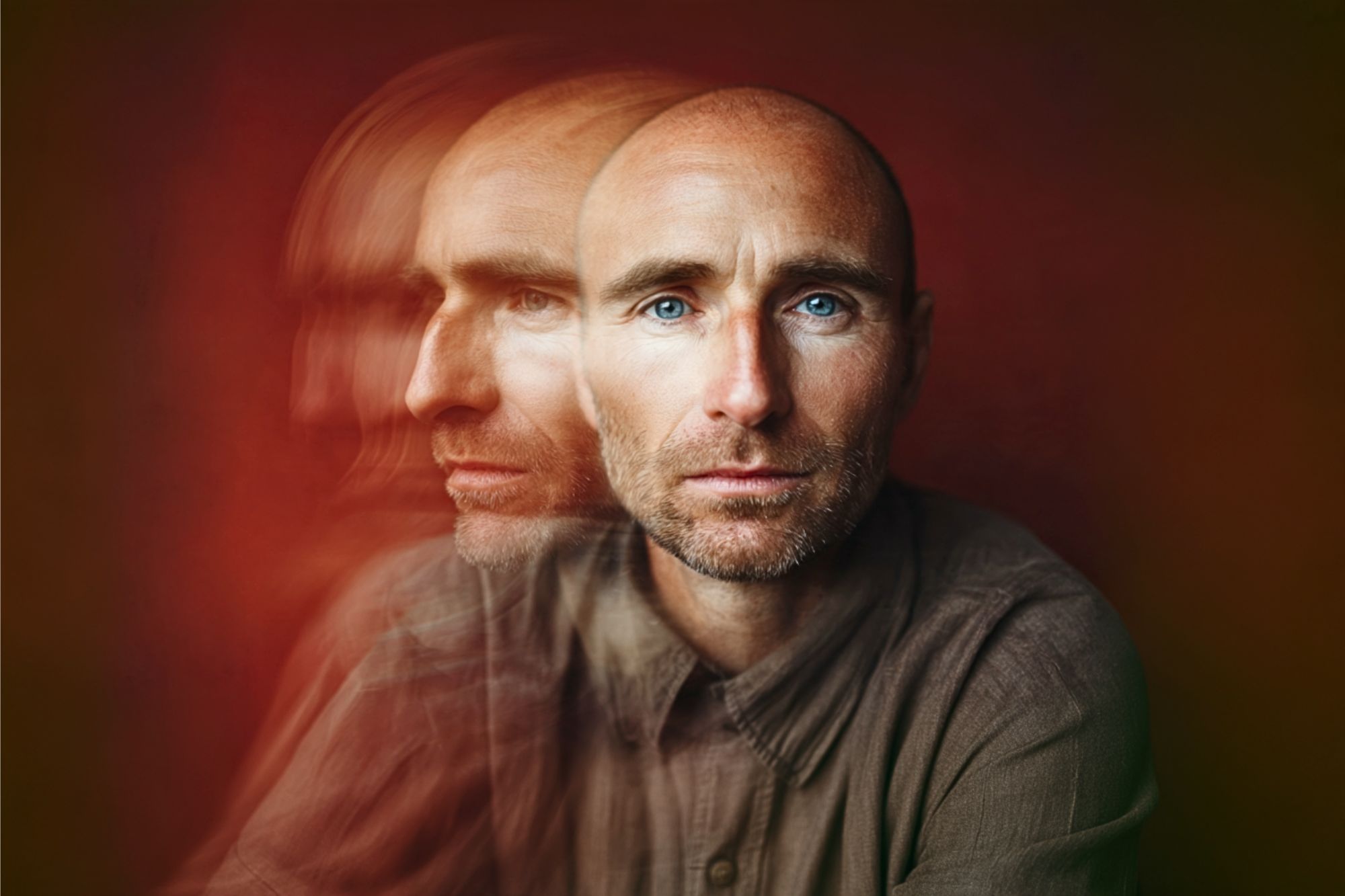 A man with blue eyes and a trimmed beard sits against a reddish background. His face appears twice—the main face in focus and a blurred duplicate to the left, creating a double exposure effect.